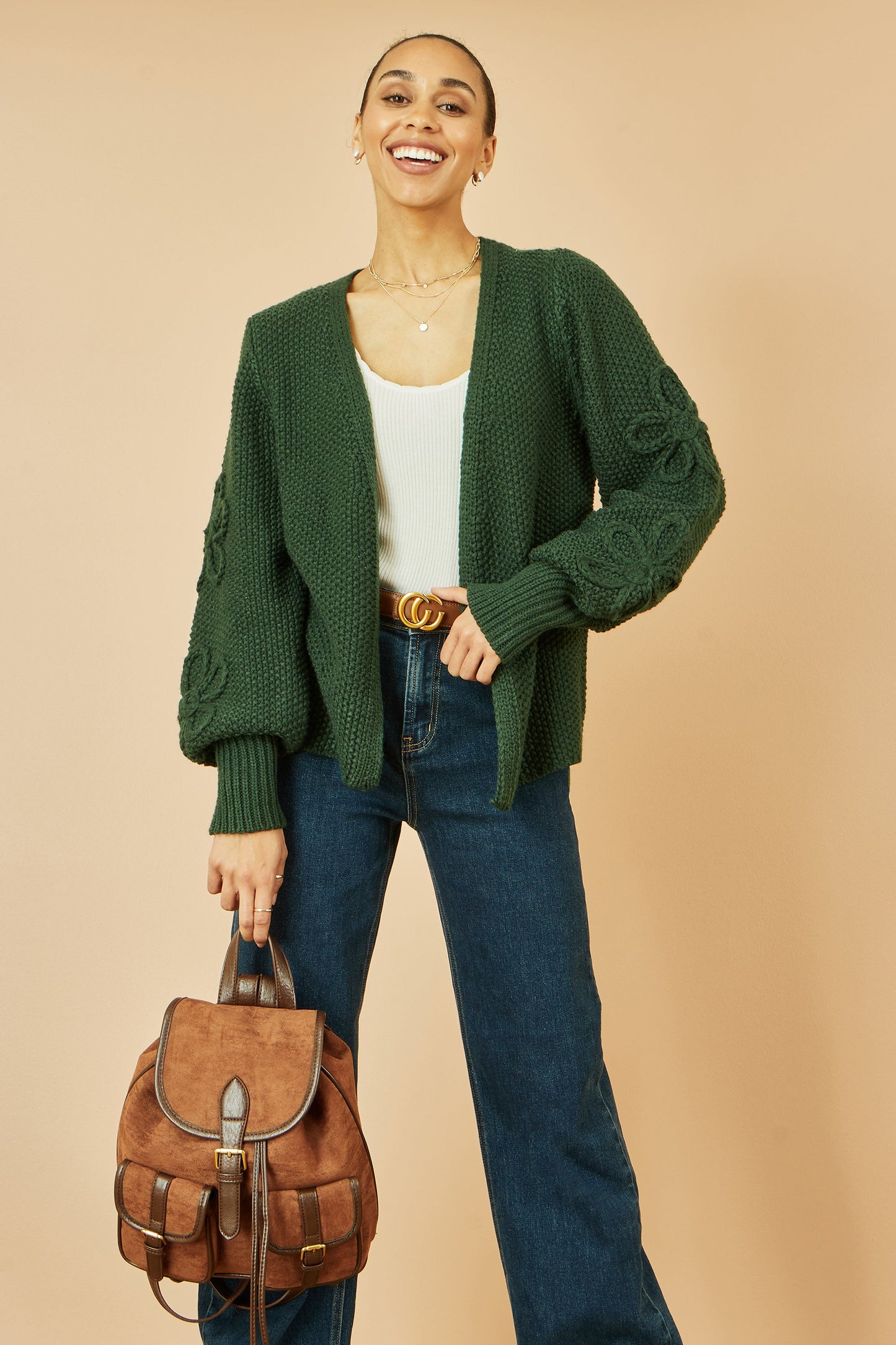 Woman wearing a green cardigan, white top, and blue jeans, holding a brown leather backpack against a beige background.