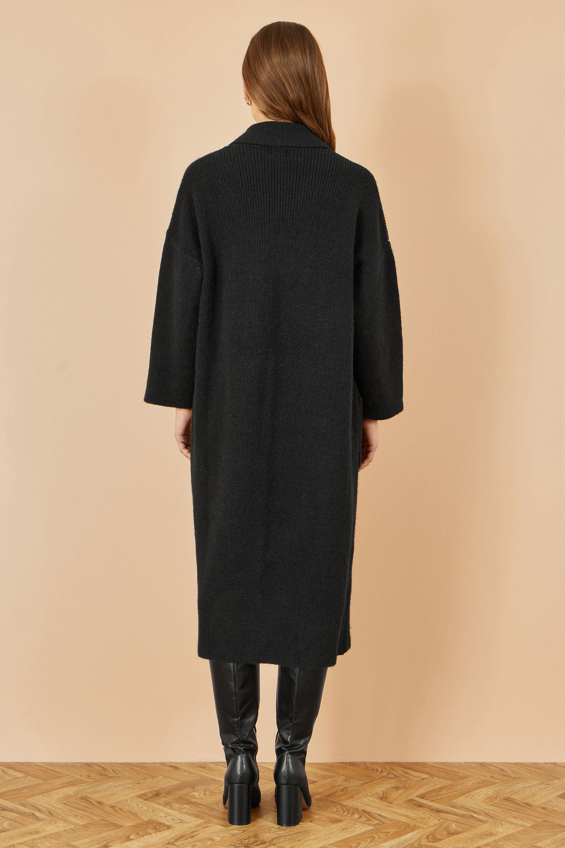 Person wearing a black coat standing against a beige wall.