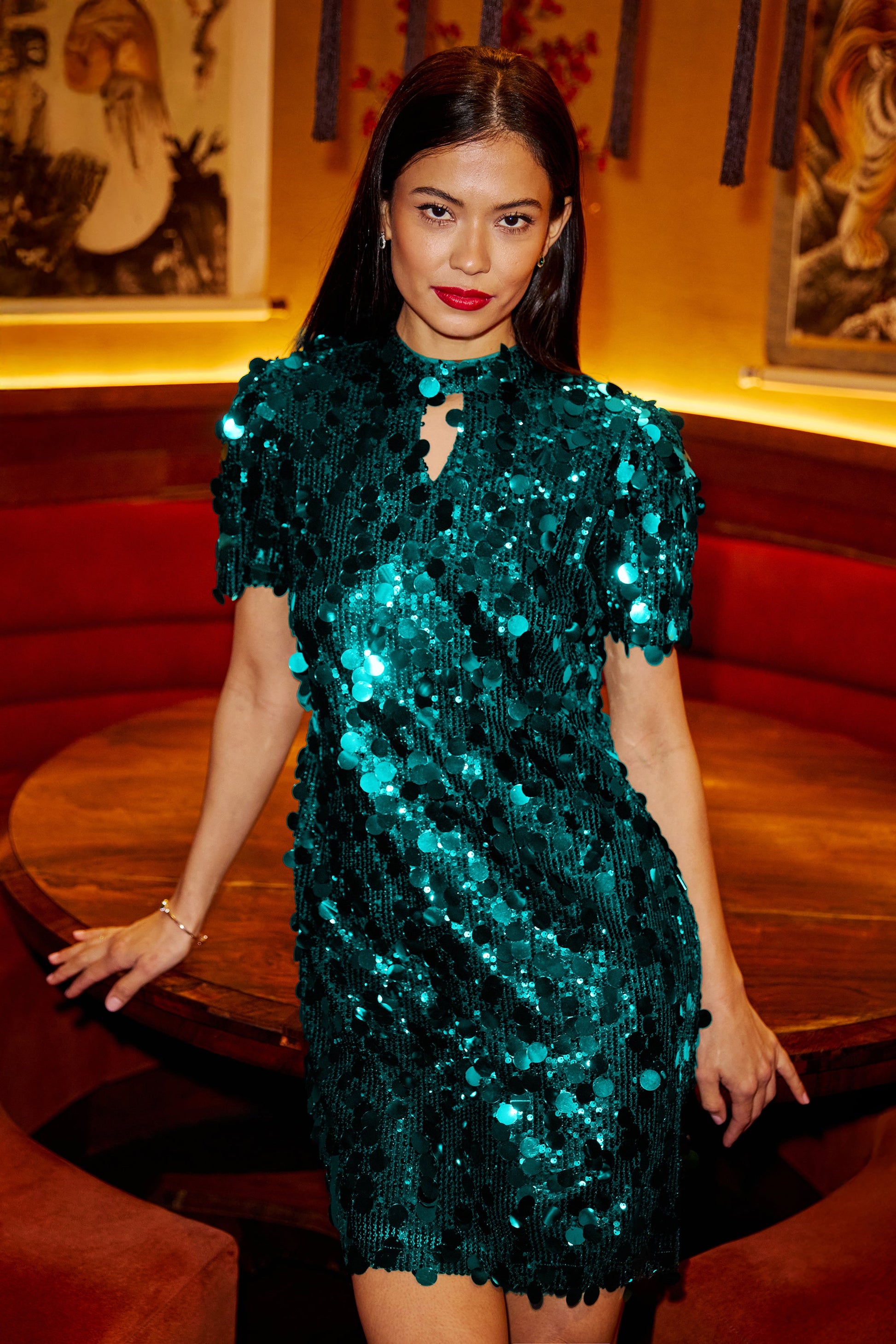 Woman wearing a green sequin dress in an indoor setting with red seats and framed pictures on the wall.