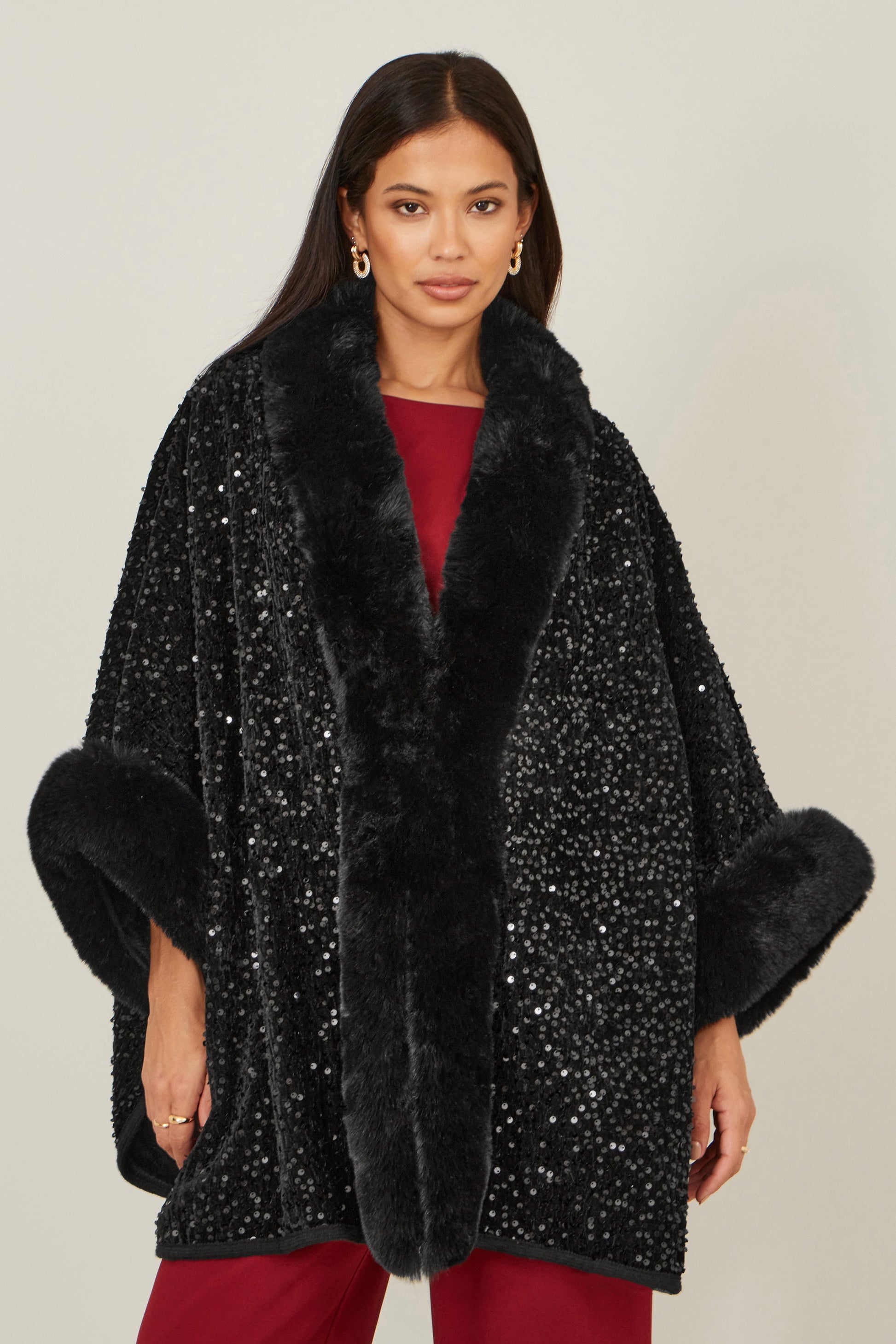 Woman wearing a black sequin cape with fur trim on a plain background