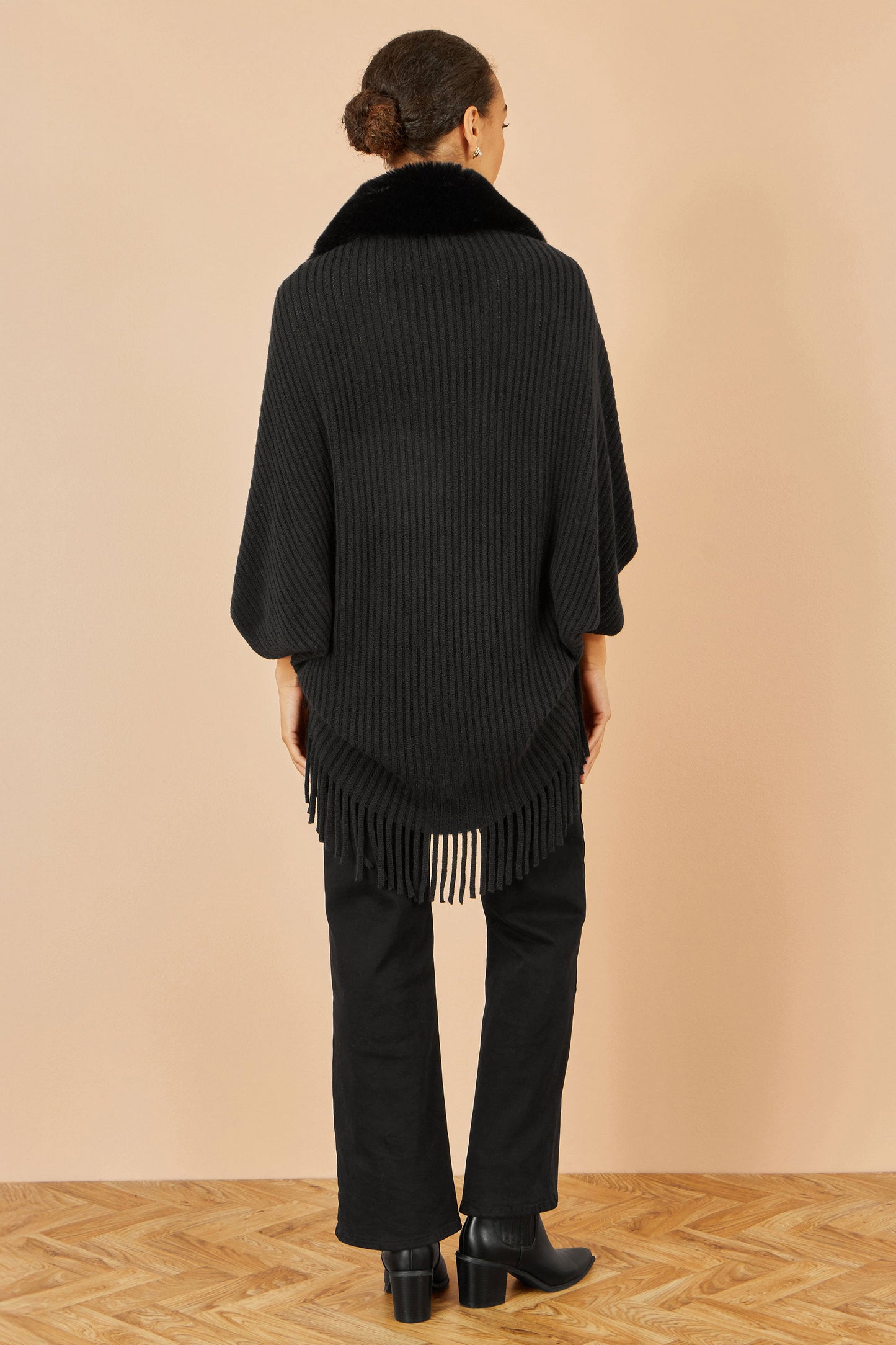 Person wearing a black knit poncho with fringe details on a beige background