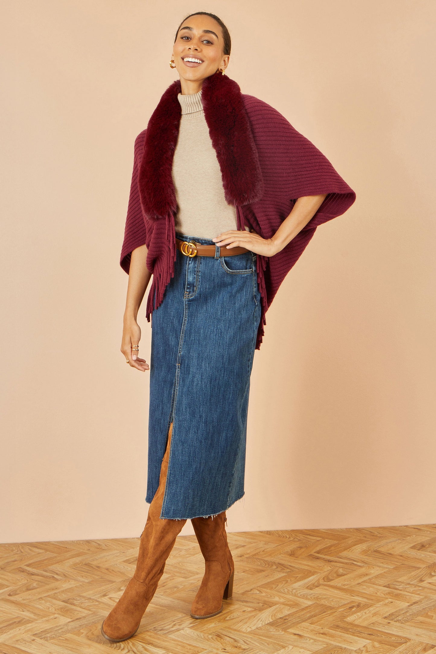 Woman wearing a burgundy poncho, beige sweater, blue denim skirt, and brown boots on a beige background