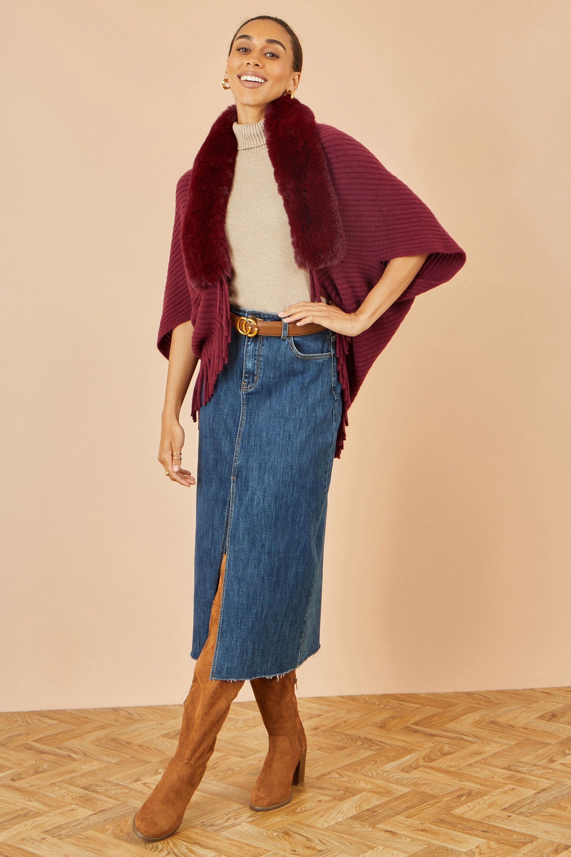 Woman wearing a burgundy poncho, beige sweater, blue denim skirt, and brown boots on a beige background