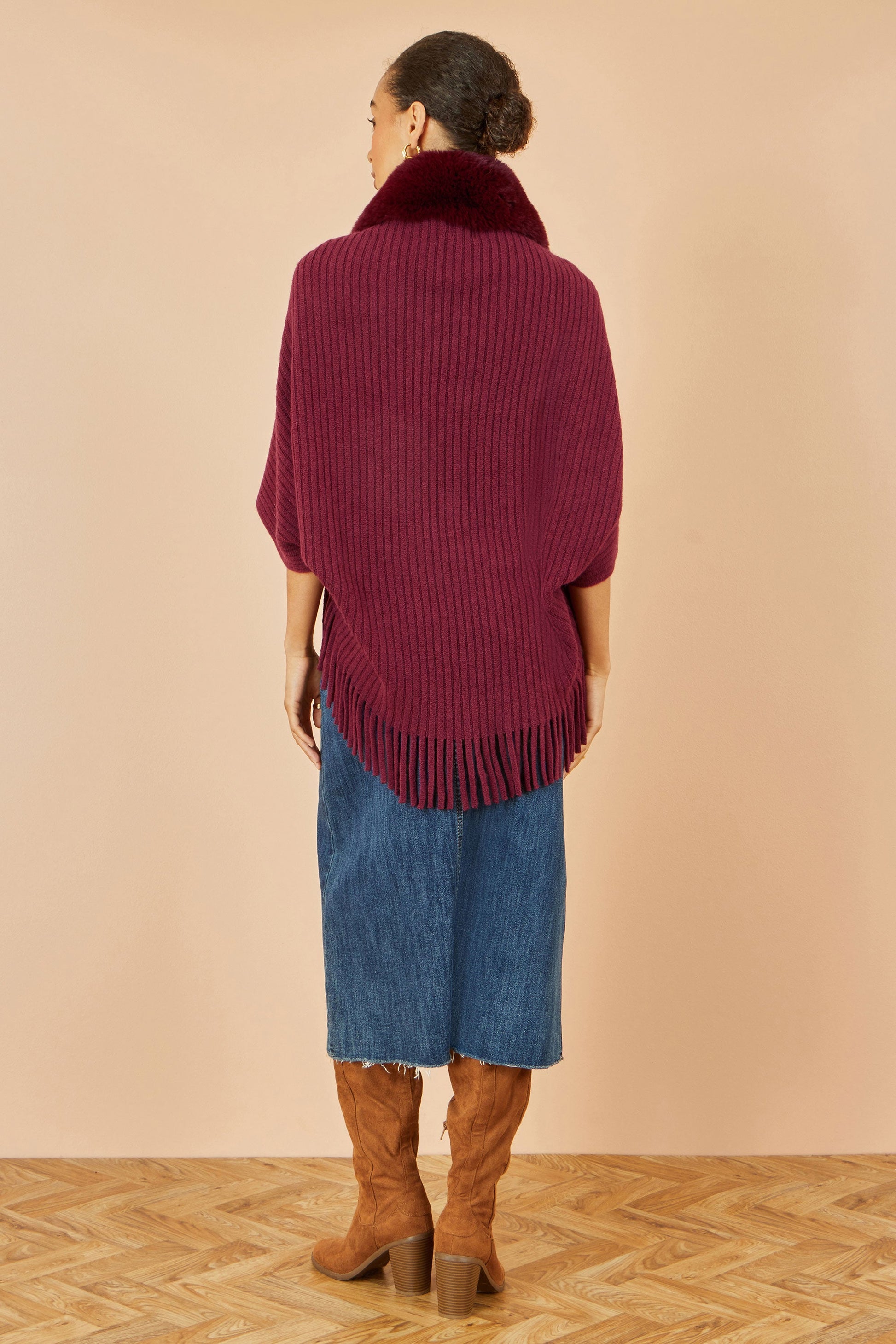 Woman wearing a burgundy fringed sweater over a blue dress with brown boots on a beige background