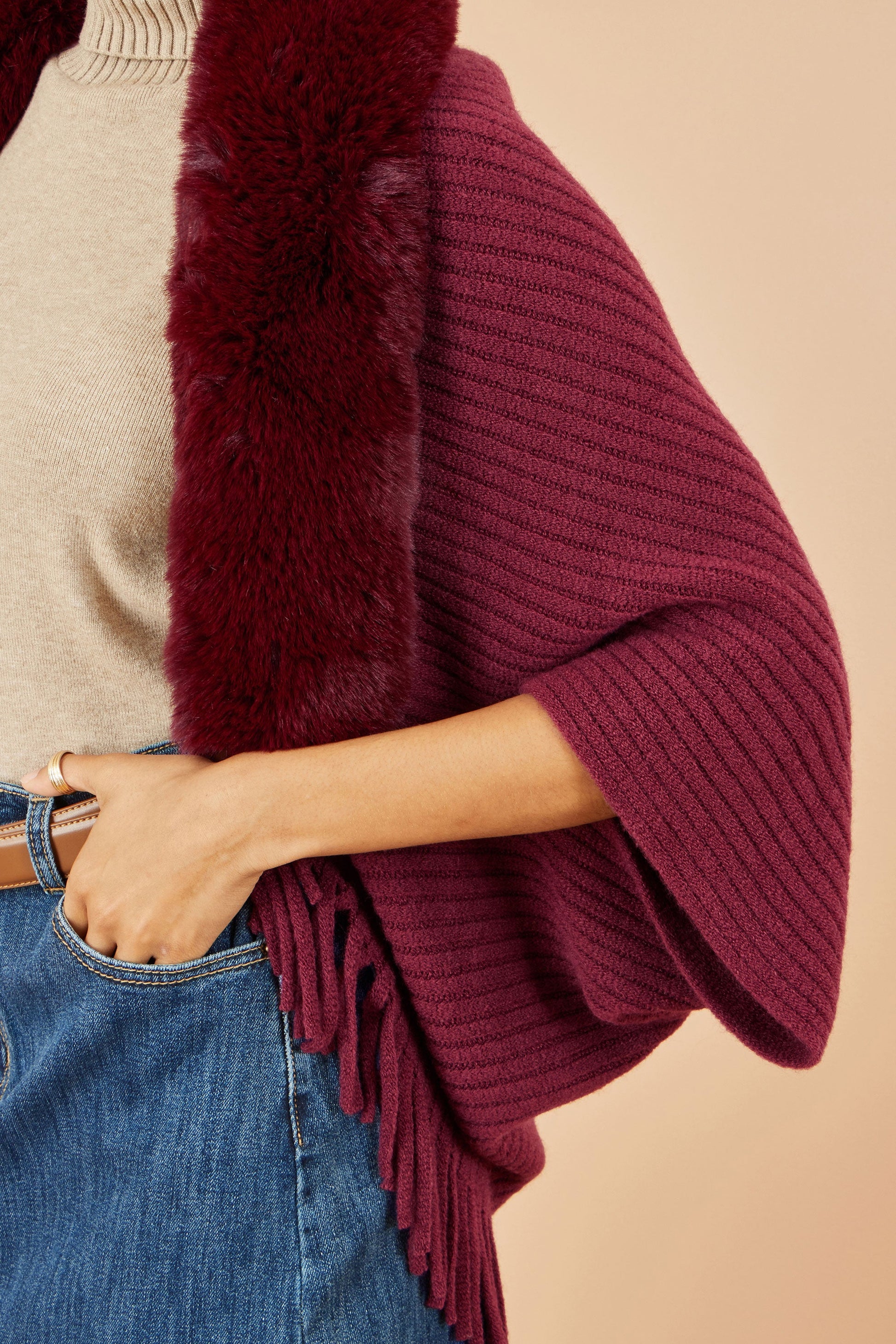 Maroon knitted poncho with fur trim worn by a person on a beige background