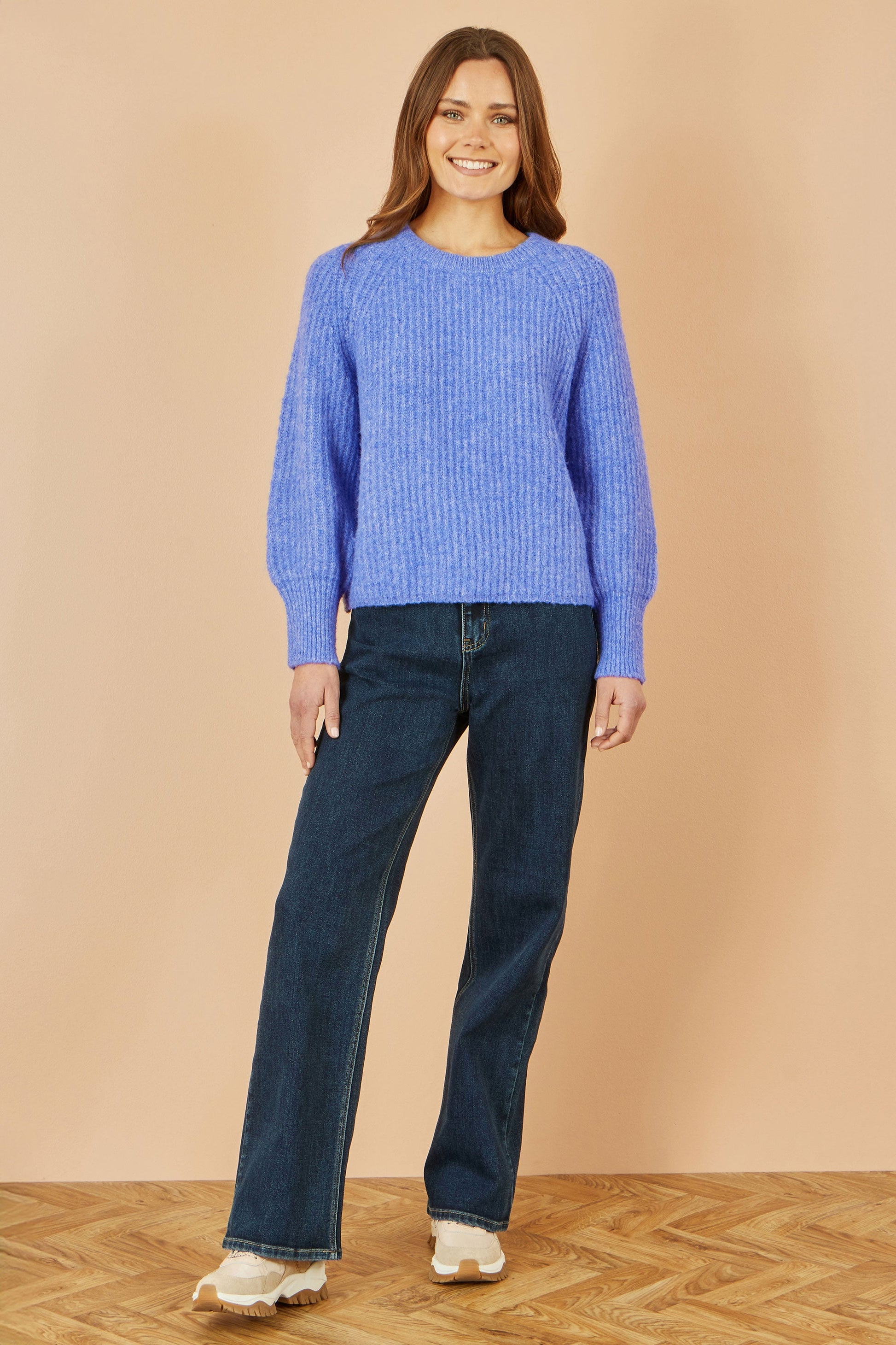 Woman wearing a blue sweater and jeans standing against a beige wall.