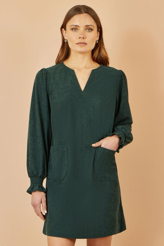 Woman wearing a green dress with long sleeves and pockets on a beige background