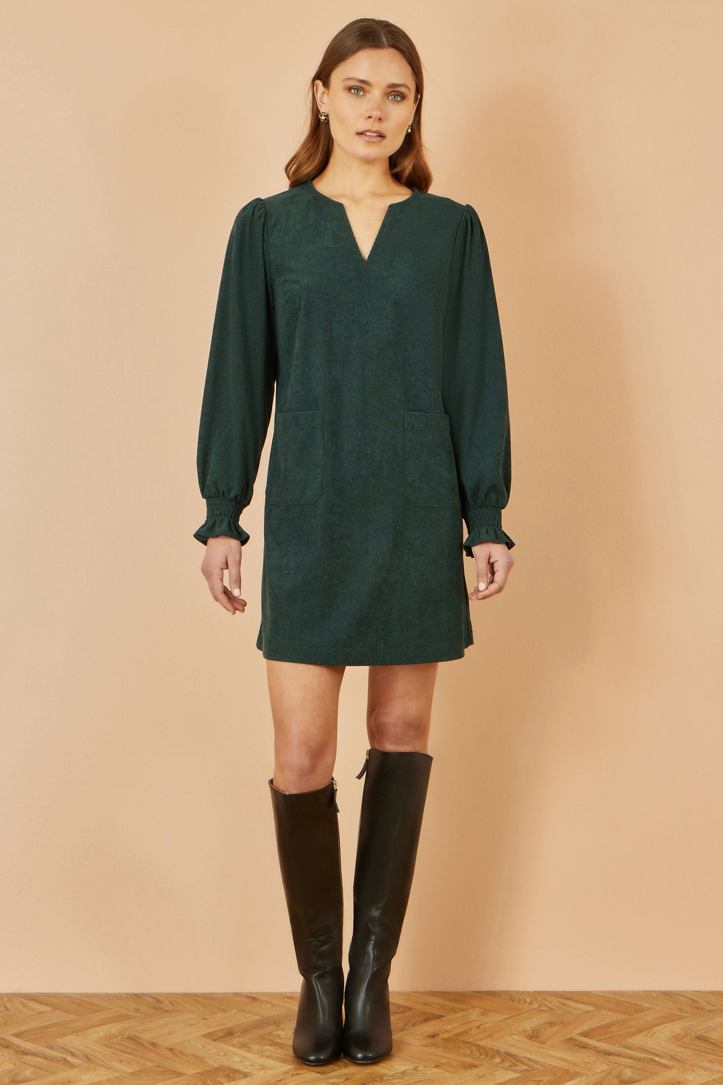 Woman wearing a green dress with long sleeves and black knee-high boots on a beige background