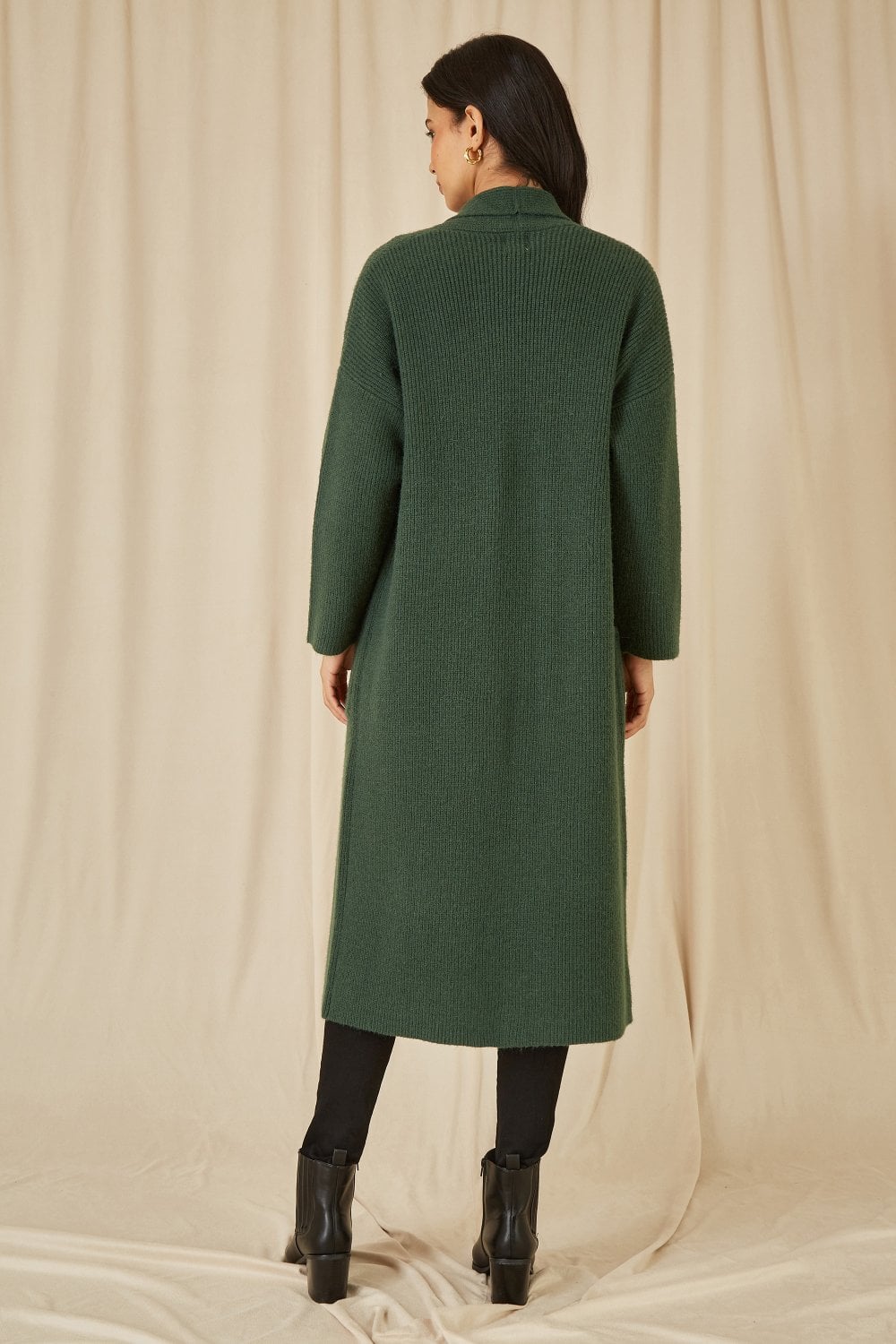 Person wearing a green coat standing against a beige curtain